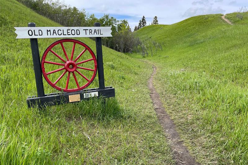 Old Macleod Trail sign with a wagon wheel beside a grassy path in Okotoks