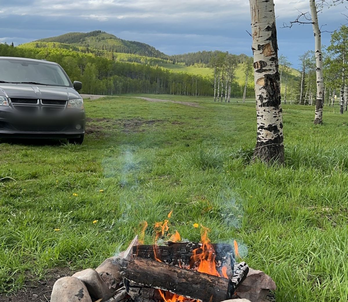 A small campfire in a stone ring on a grassy meadow with aspen trees and green hills behind