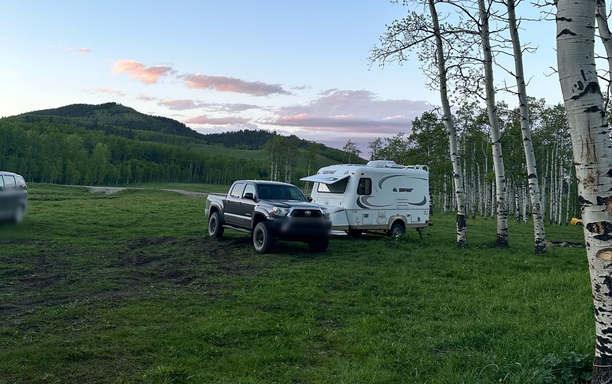 A truck and small travel trailer in a grassy clearing beside aspens at sunset